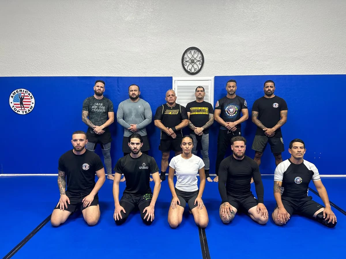 Adult no-gi class in rash guards and training shirts, one row kneeling and one standing, in front of an American Top Team wall logo