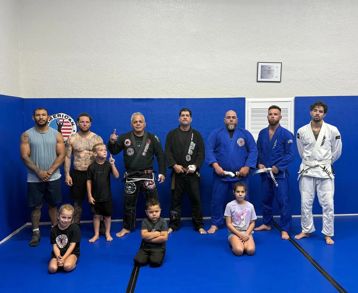 Kids and adult students in gis and training clothes posing together on blue mats in front of an American Top Team wall logo