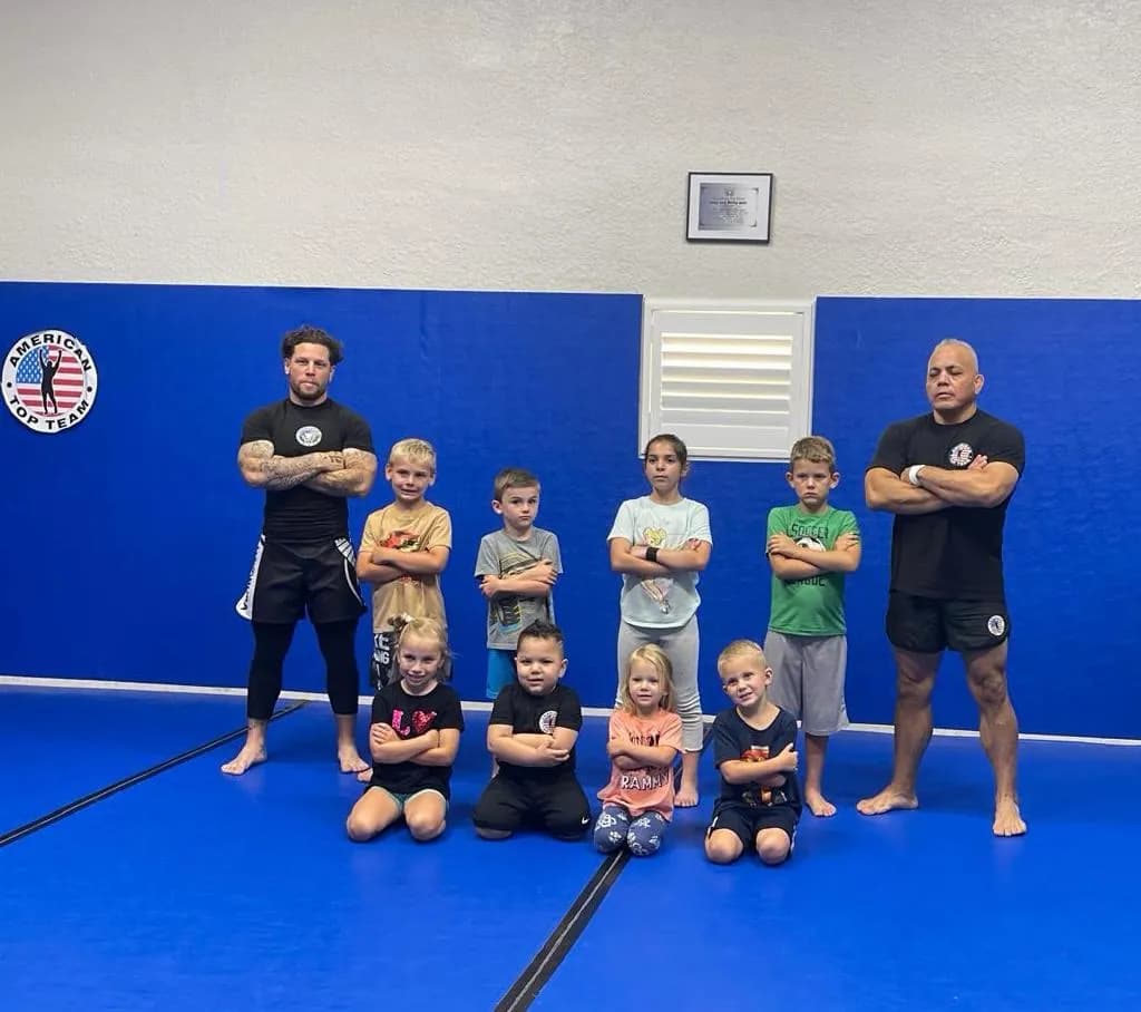 Group of young boys and girls posing with arms crossed on blue mats between two coaches, with an American Top Team wall logo