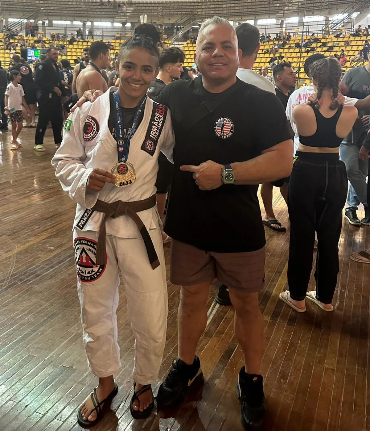 Female jiu-jitsu competitor in a white gi with a brown belt holding up a medal next to Master Willy Hardoy at a tournament venue