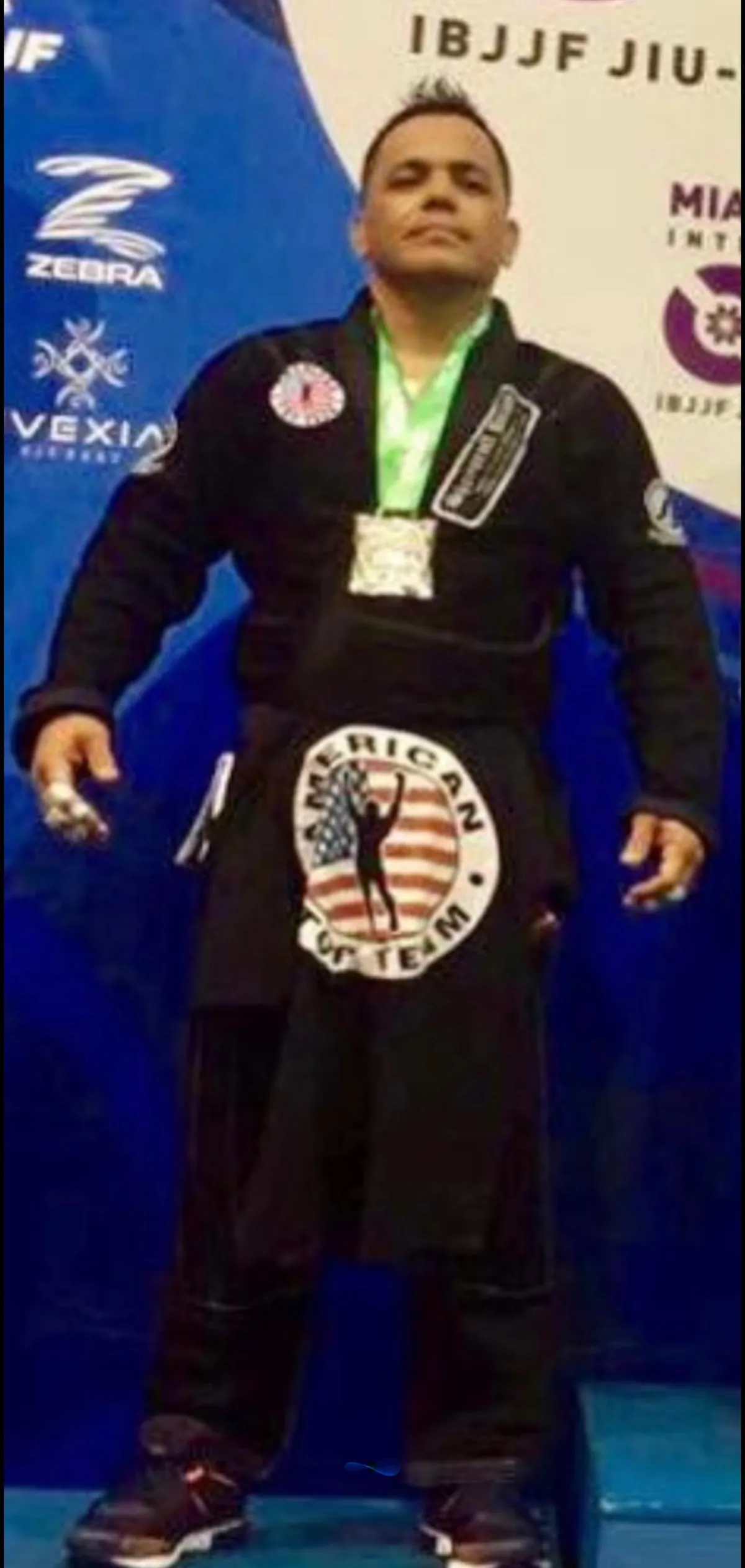 Master Willy Hardoy in a black gi with a large American Top Team patch, wearing a medal in front of an IBJJF jiu-jitsu banner