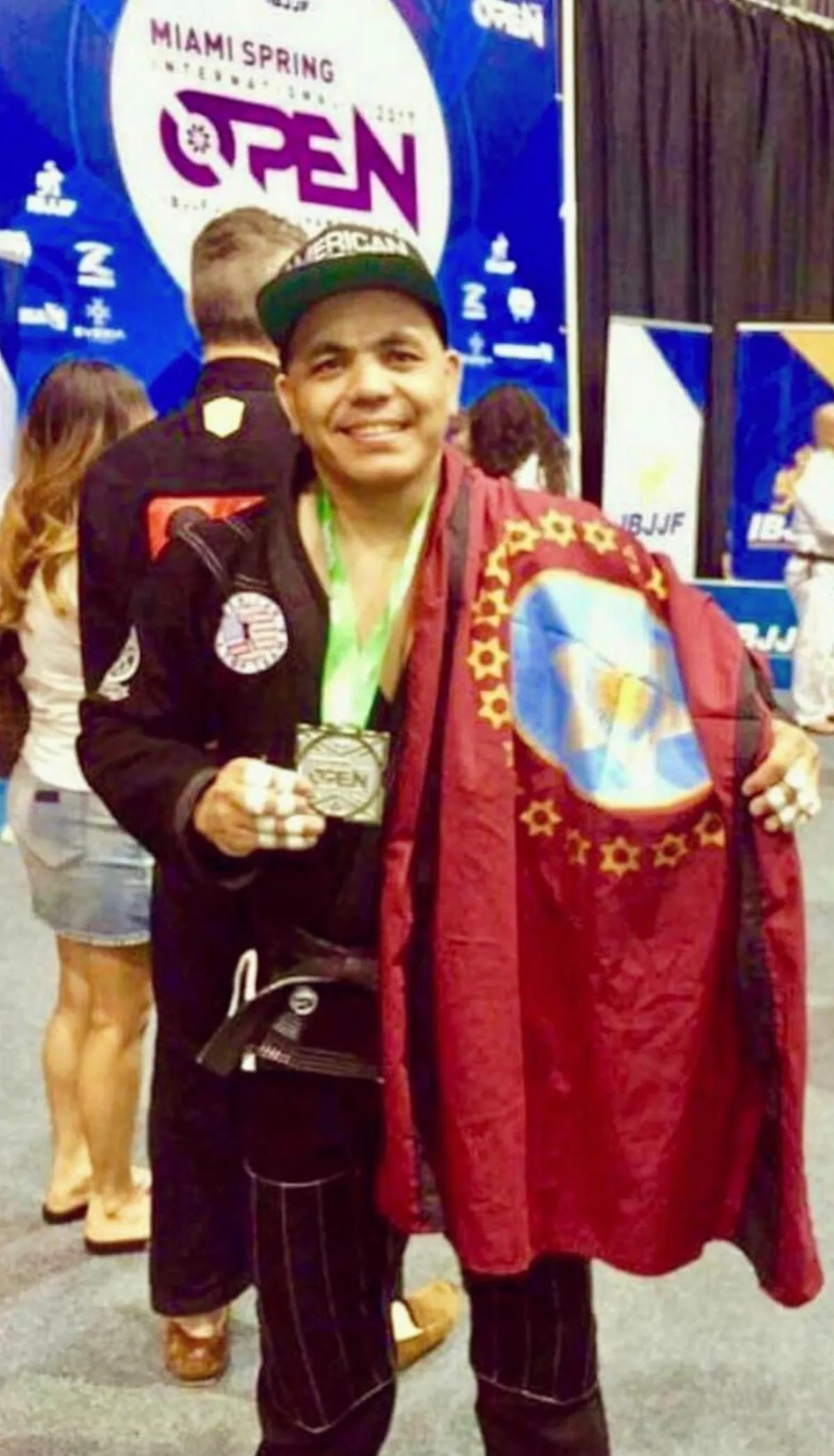 Master Willy Hardoy in a black American Top Team gi and cap, wearing a medal and draped in a maroon flag in front of a Miami Spring International Open banner