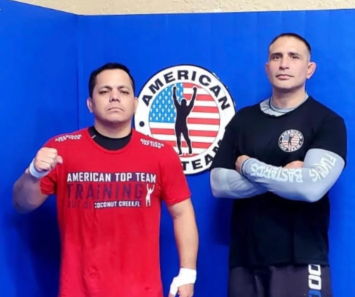 Master Willy Hardoy with black hair, wearing a red American Top Team training shirt, standing beside a teammate in front of a large American Top Team logo