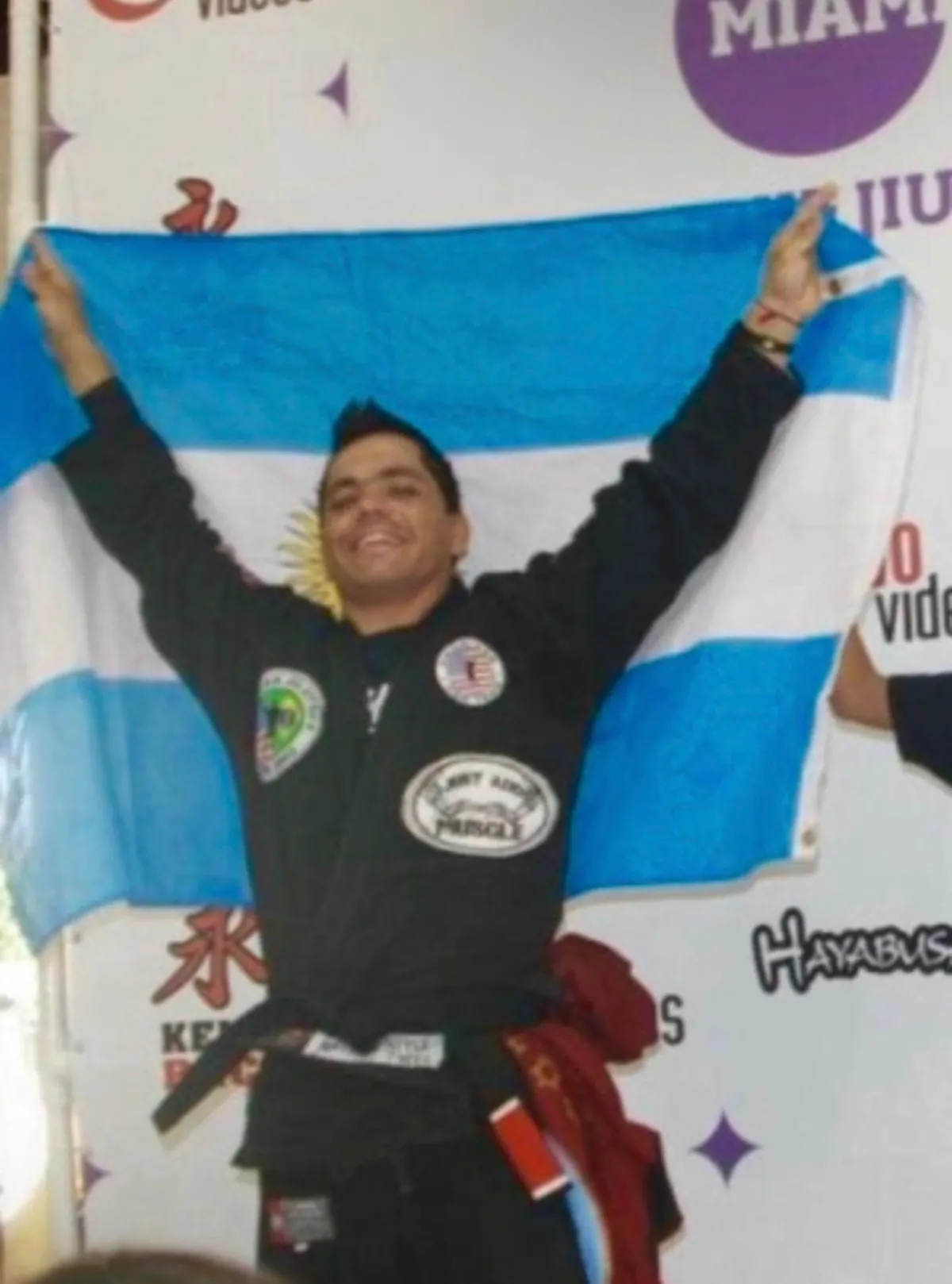 Master Willy Hardoy with black hair in a black gi, holding a light blue and white flag above his head in front of a tournament backdrop