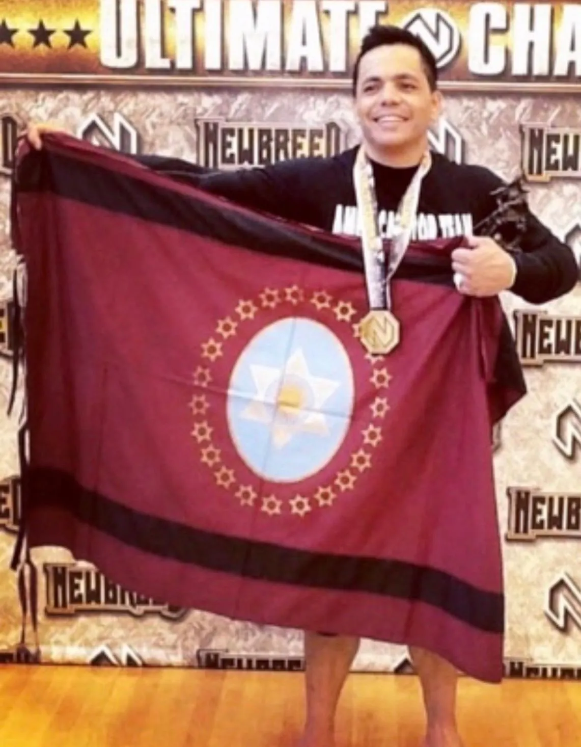 Master Willy Hardoy with black hair wearing a medal and holding a maroon flag in front of a Newbreed Ultimate Challenge backdrop