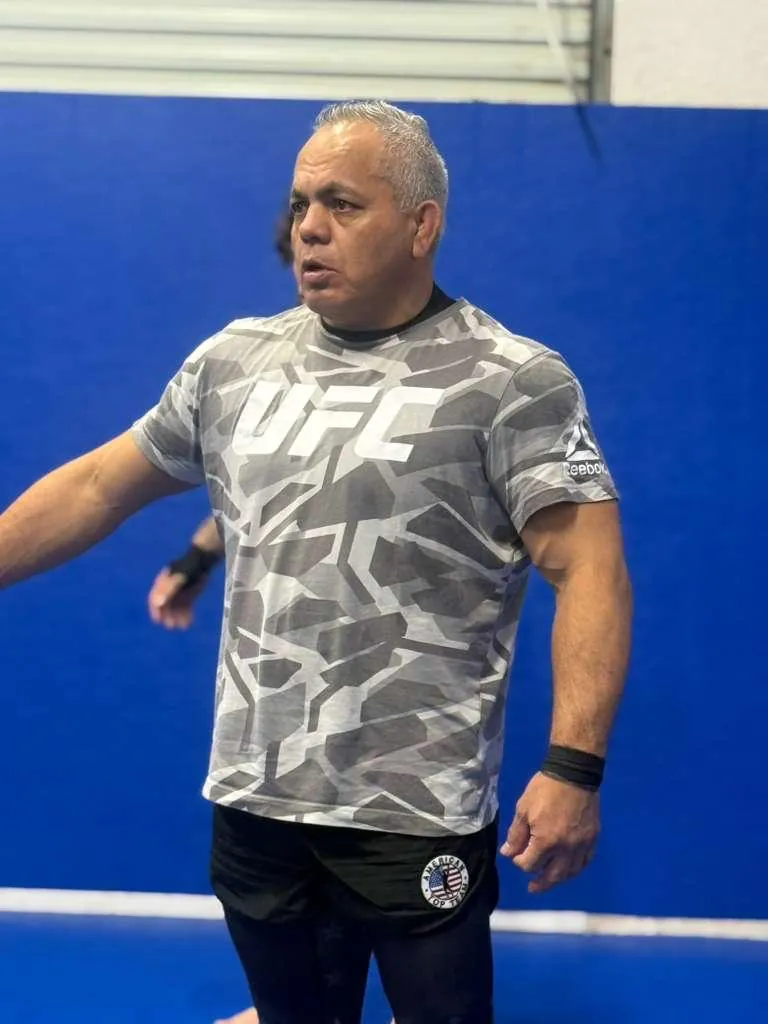 Portrait of Master Willy Hardoy today, with white hair, in a gray camo T-shirt, gesturing while teaching on blue mats