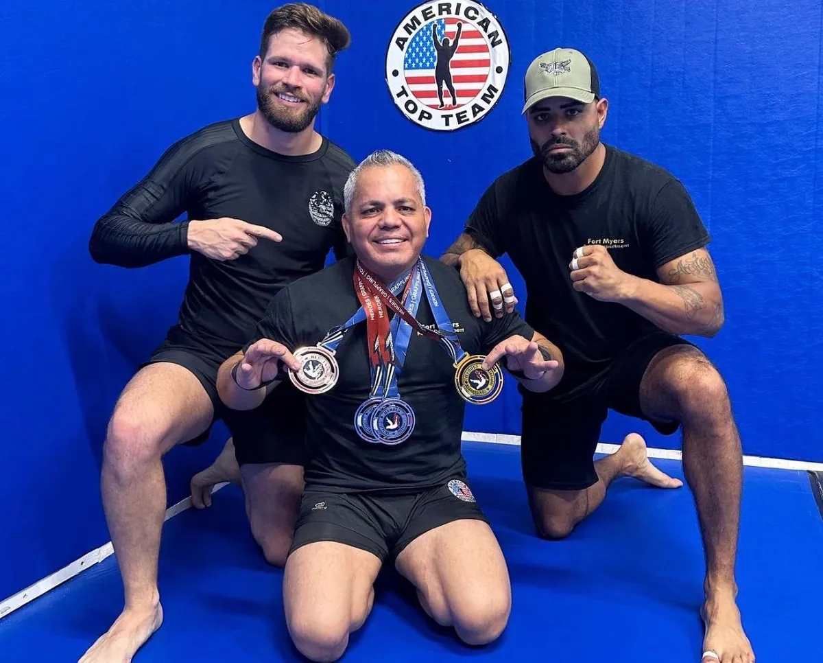 Master Willy Hardoy with white hair kneeling on blue mats holding several medals, flanked by two students in front of an American Top Team logo